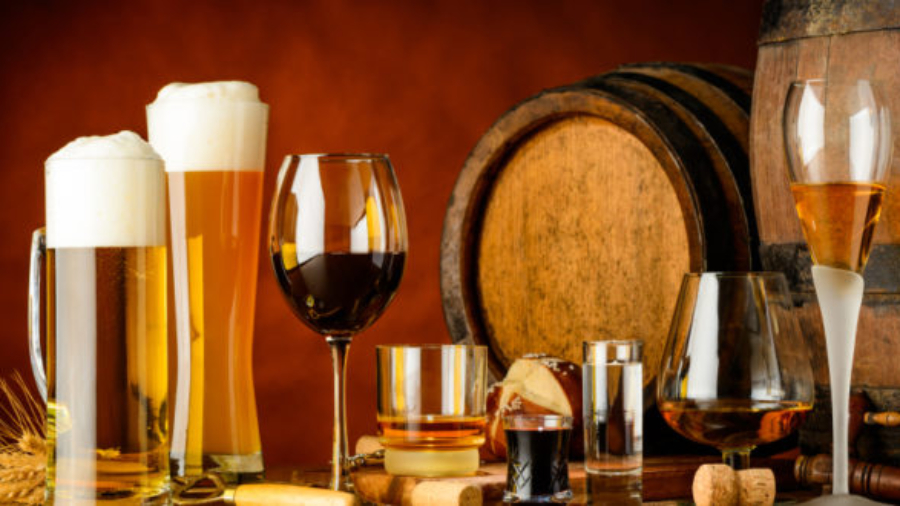 alcoholic drinks on wooden table in glasses, mugs and shots with barrel in background